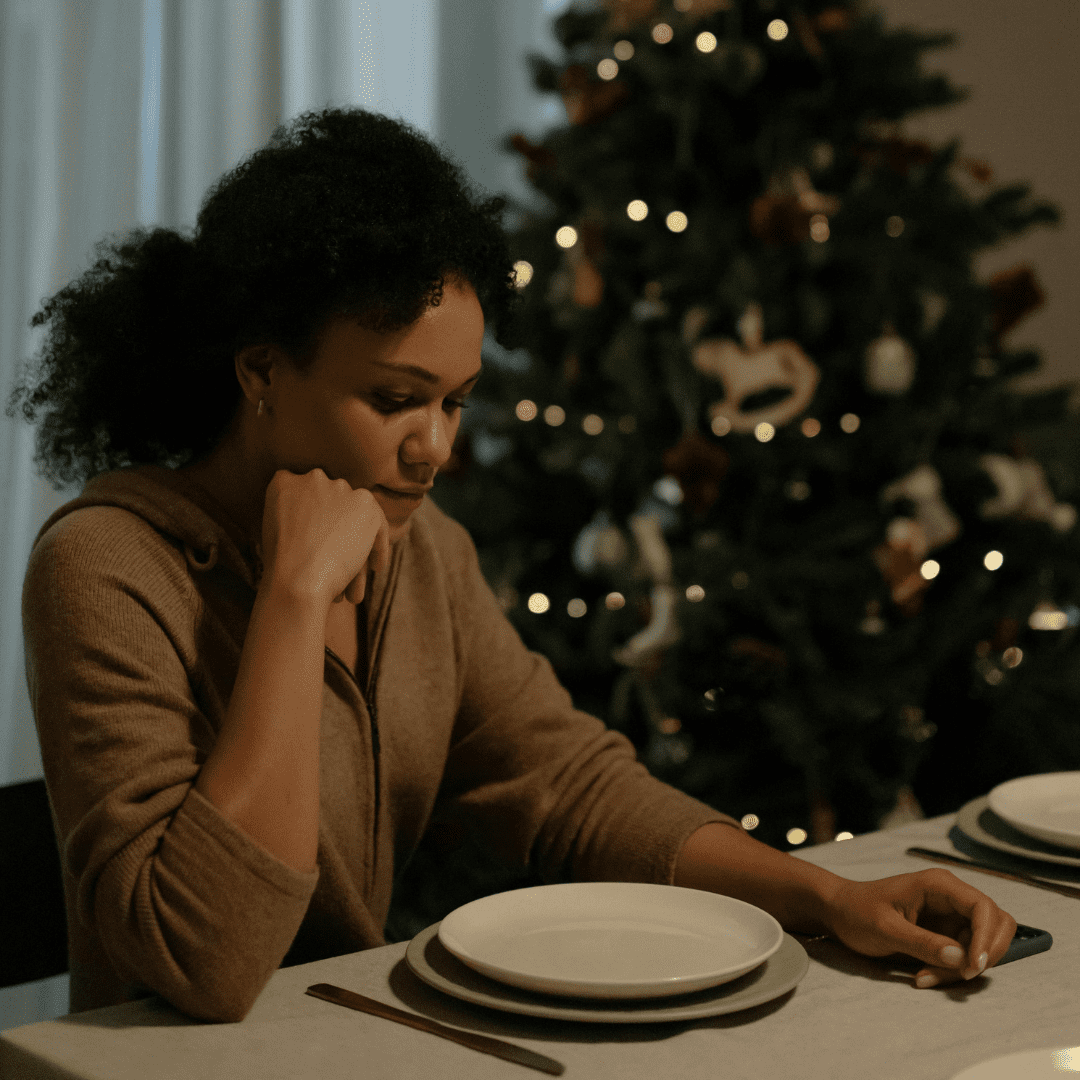 sad woman sitting with hand on chin at dining table next to Christmas tree