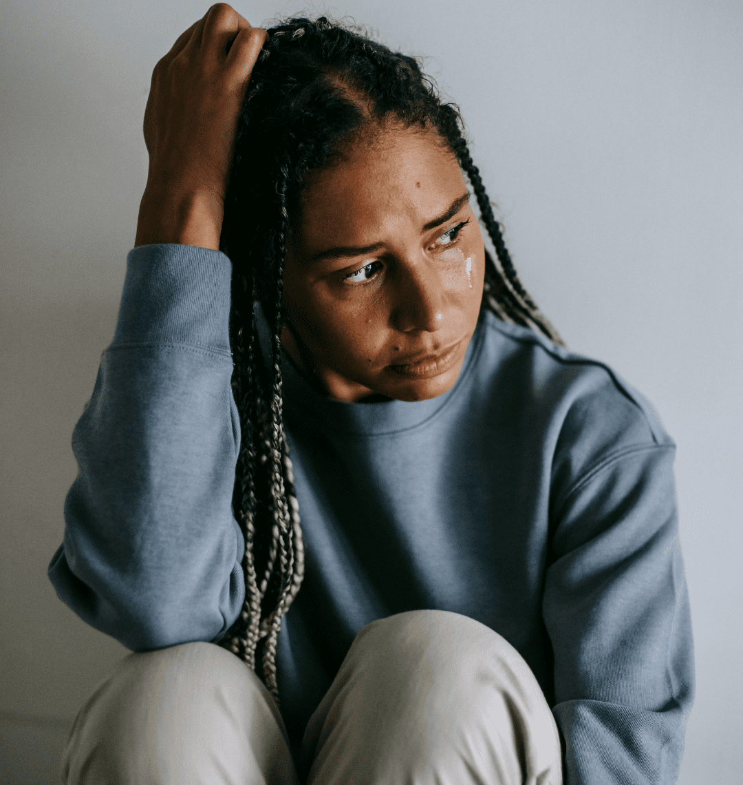 A young woman with braided hair, wearing a gray sweater, sitting with her hand on her head, tears running down her face.