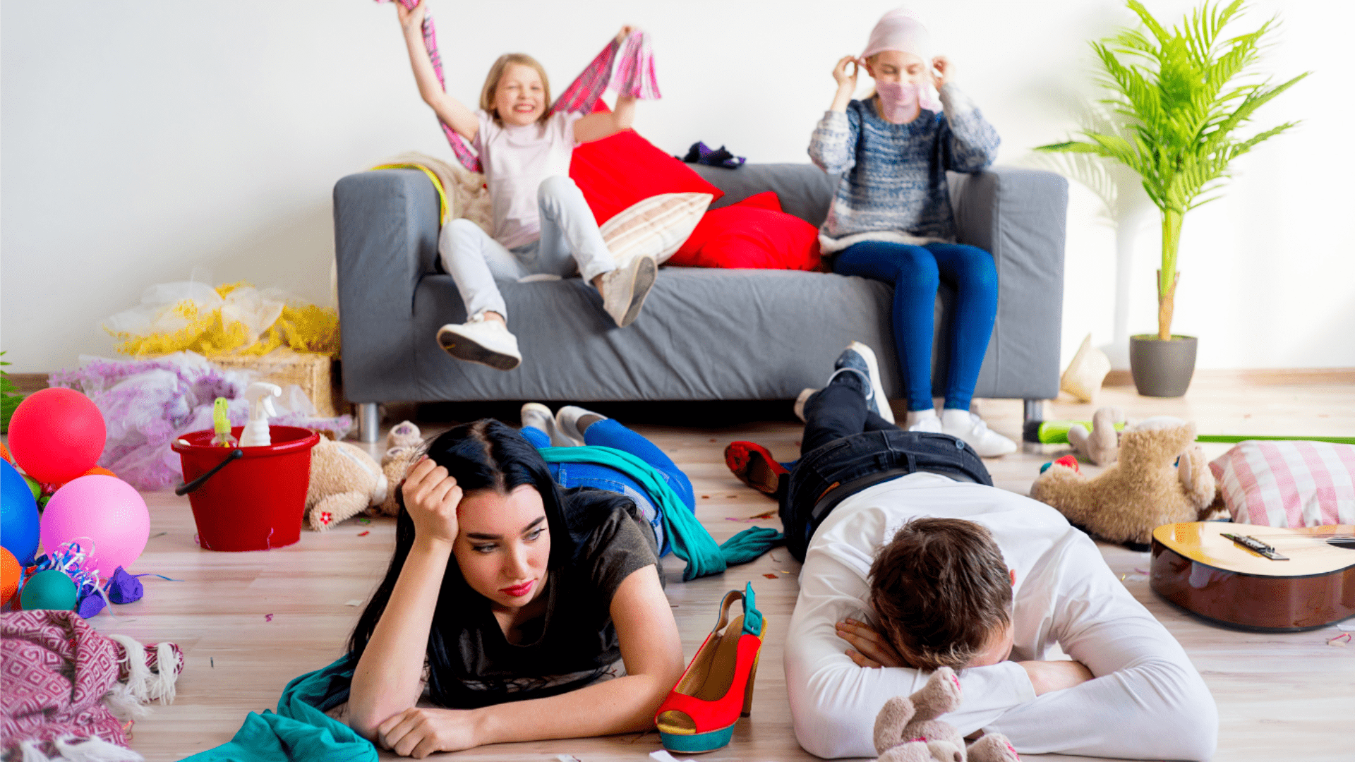 Exhausted parents lay on the floor of a messy living room, surrounded by toys, balloons, and scattered decorations, while children continue to play energetically in the background.