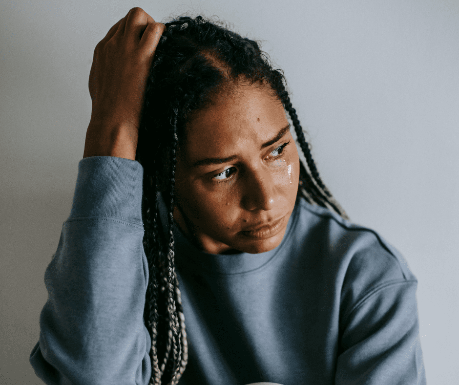 A young woman with braided hair, wearing a gray sweater, sitting with her hand on her head, tears running down her face.