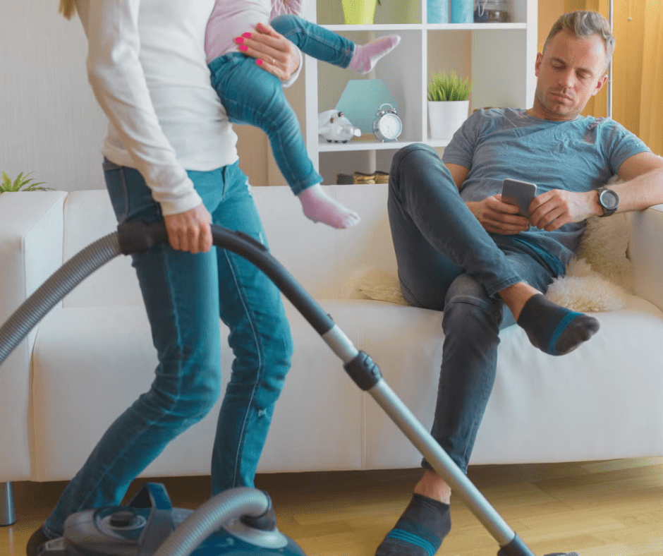 A woman vacuums while holding a child, while a man sits on the couch looking at his phone, appearing disengaged, highlighting an imbalance in household responsibilities.