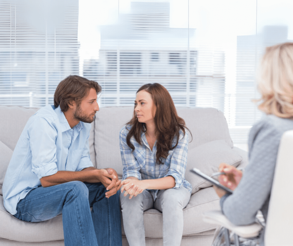 A couple sits closely together on a couch in a therapist’s office, engaged in a serious conversation, reflecting their desire to strengthen their relationship before problems arise