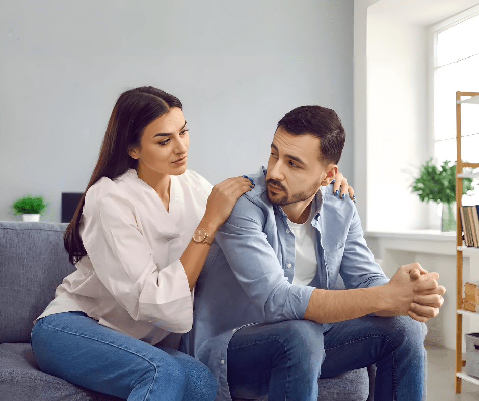 A concerned woman sits beside her distant partner, gently placing a hand on his shoulder as he looks away, deep in thought. The atmosphere is quiet and introspective.