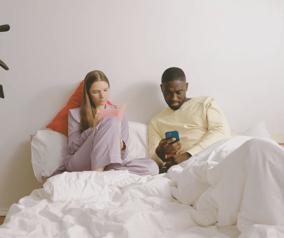 A couple sits in bed together, but they seem disconnected—she is focused on reading a book while he scrolls through his phone, both absorbed in their own worlds