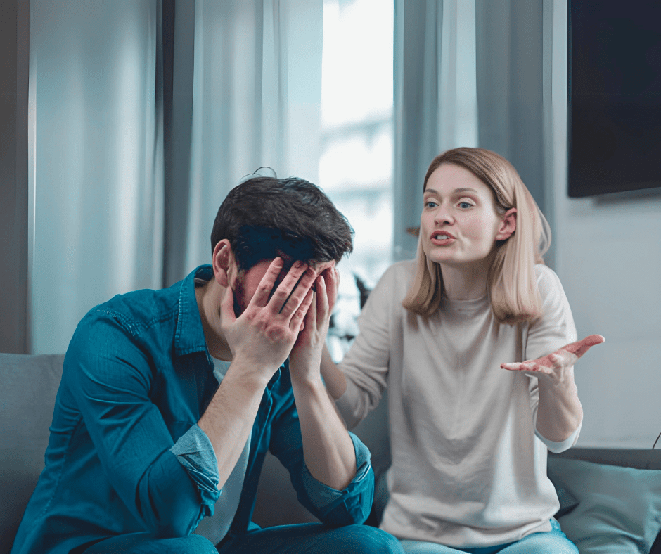 A frustrated woman gestures while speaking to a man who sits with his face in his hands, appearing overwhelmed and disengaged, reflecting a tense argument