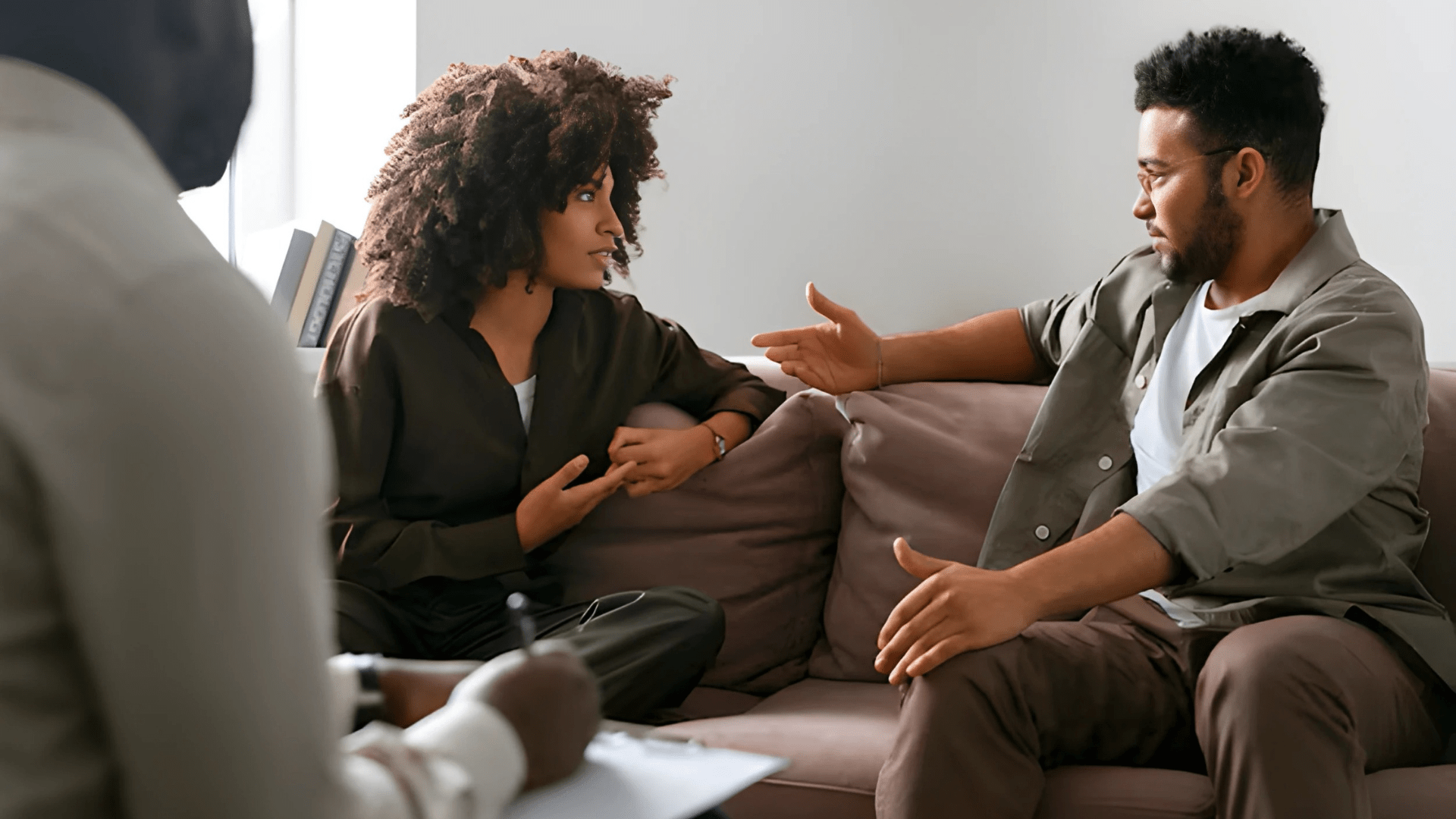 A woman and a man sit on a couch engaged in a serious discussion, while a therapist takes notes in the foreground