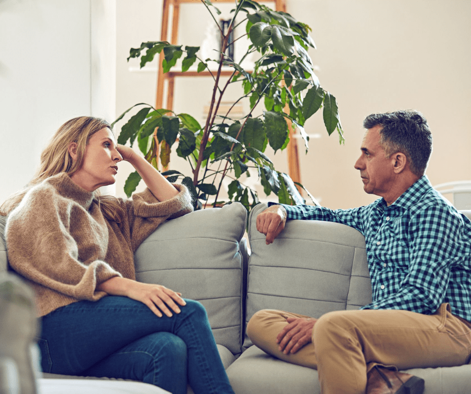 A woman in a brown sweater and a man in a checkered shirt sit on a couch, engaged in a deep discussion in a well-lit living room