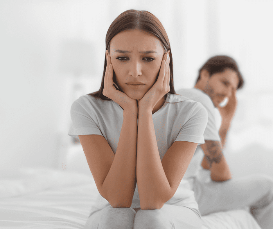 A sad woman sits on the bed, while her frustrated partner sits behind her, both distant