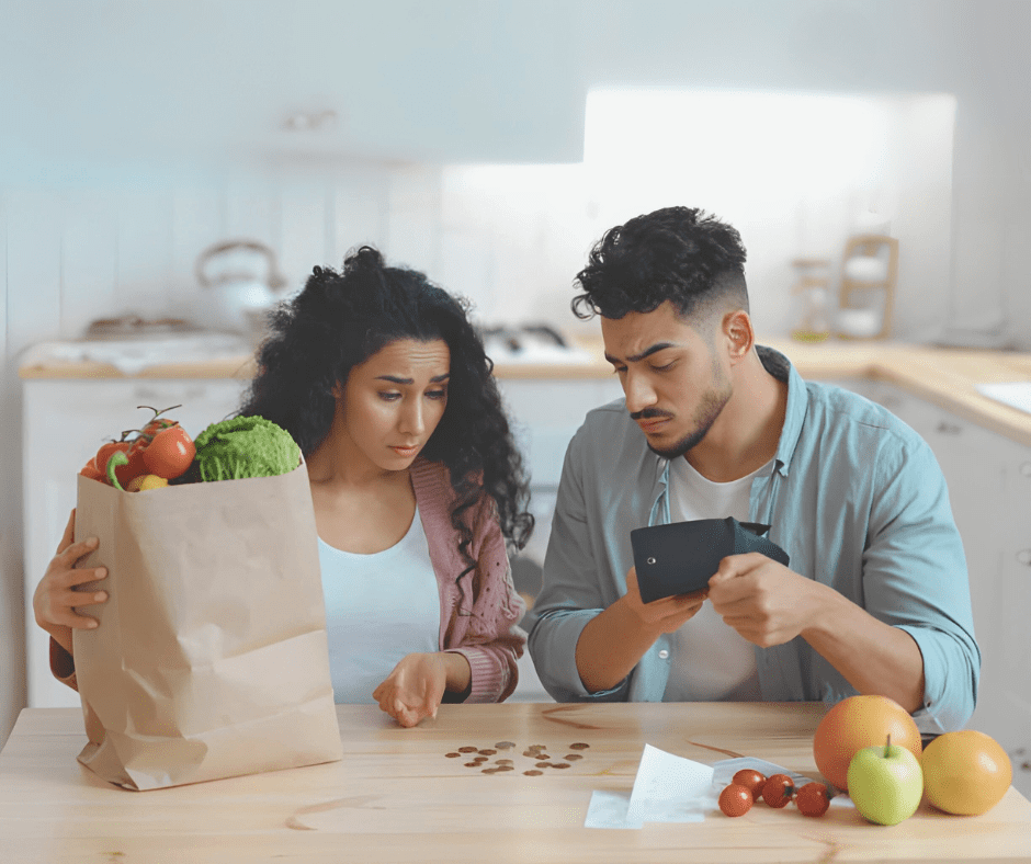 A worried couple sits at a kitchen table, counting their last coins. The woman holds a paper grocery bag with fresh produce, while the man checks his empty wallet, looking concerned.