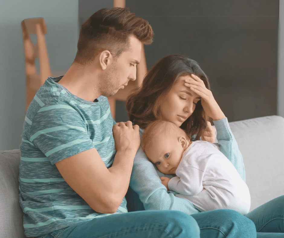 A tired mother cradles her baby while resting her head in her hand, looking overwhelmed. Her partner sits beside her, appearing concerned yet unsure how to help.
