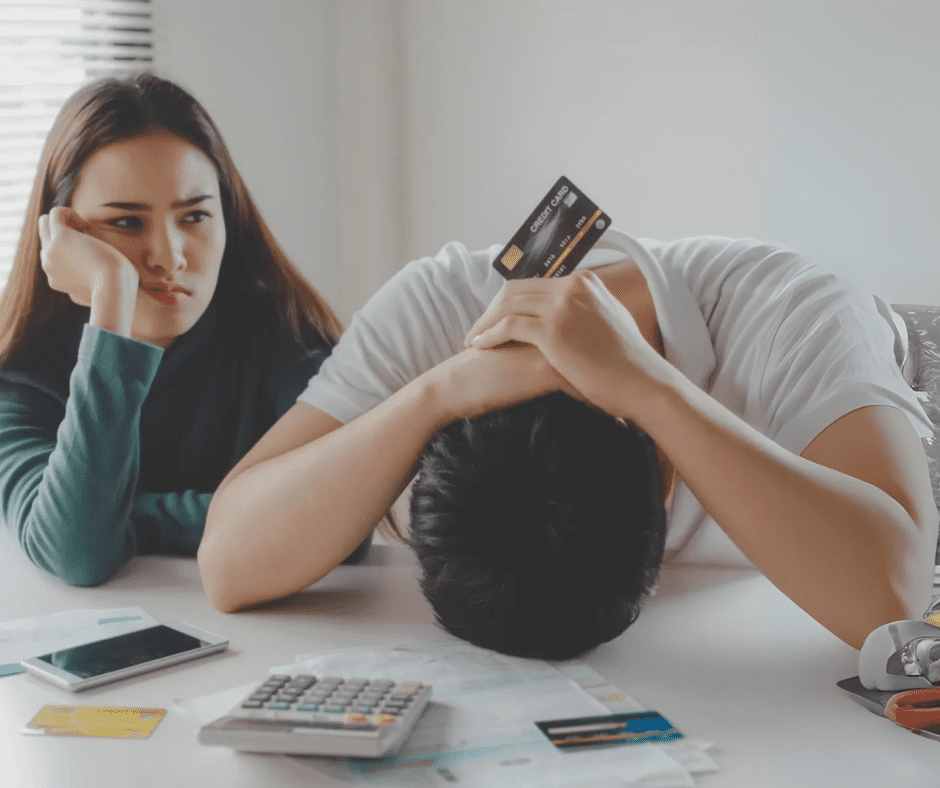 A stressed man buries his head in his arms, holding a credit card, while his frustrated partner glares at him over a pile of bills.