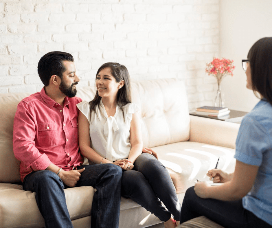 A smiling couple sits together on a couch in a therapist’s office, looking at each other warmly, symbolizing trust, connection, and mutual understanding
