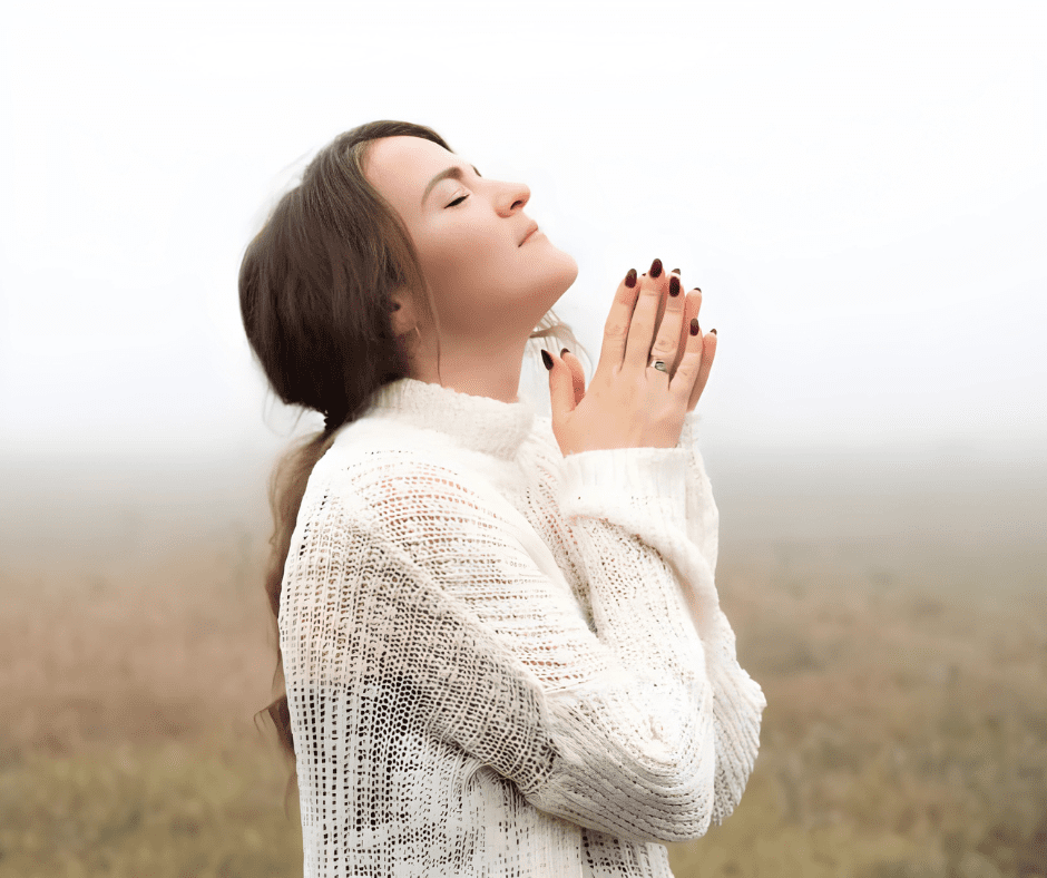 Peaceful young woman with eyes closed and hands together in prayer, standing in a foggy field wearing a white sweater.