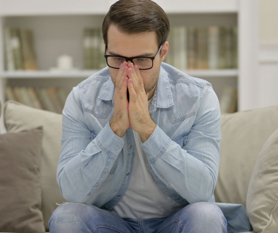 Young man covering her face with both hands, sitting by a window and expressing feelings of stress or overwhelm