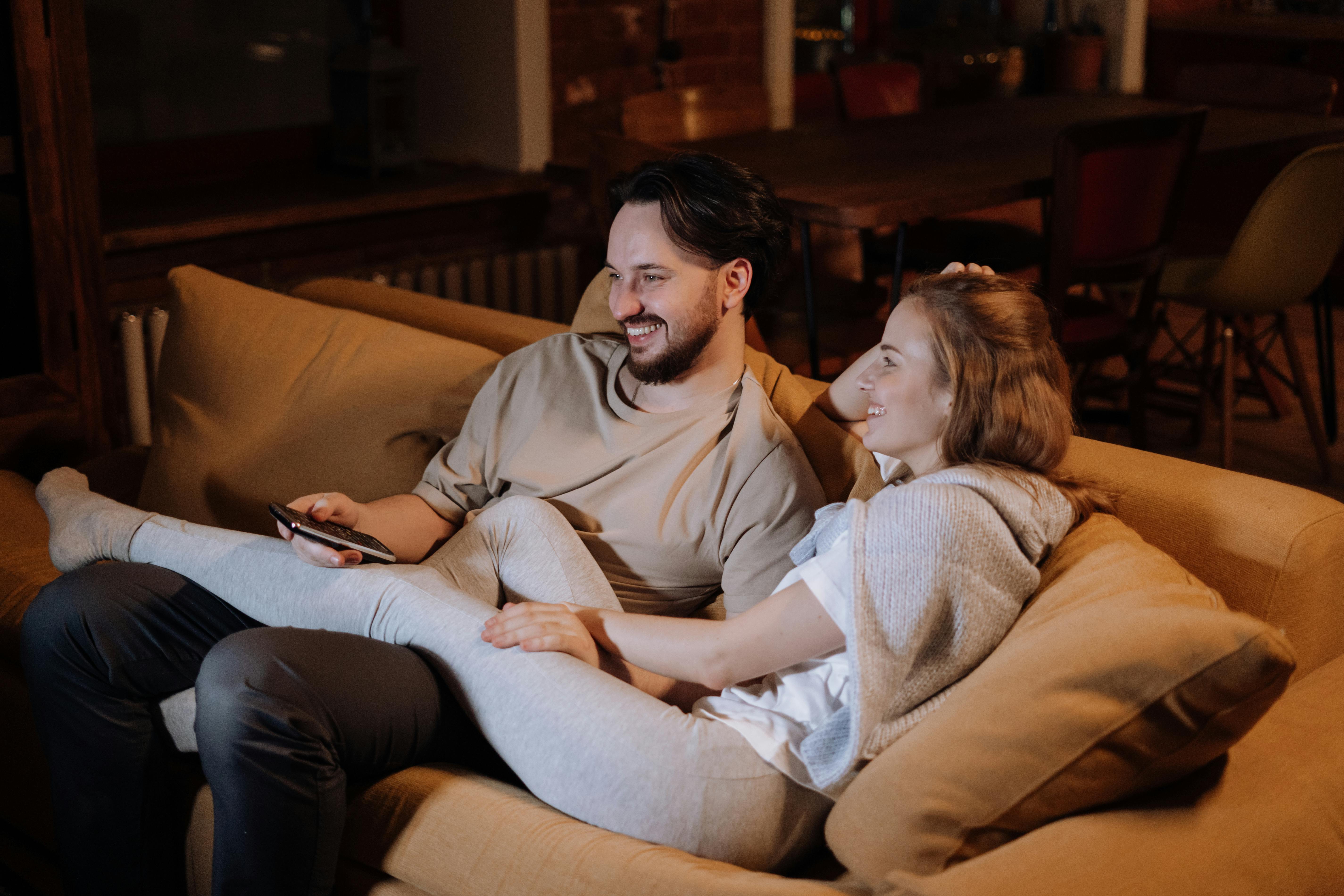 A smiling engaged couple relaxing together on a cozy couch at home, watching TV and enjoying quality time.